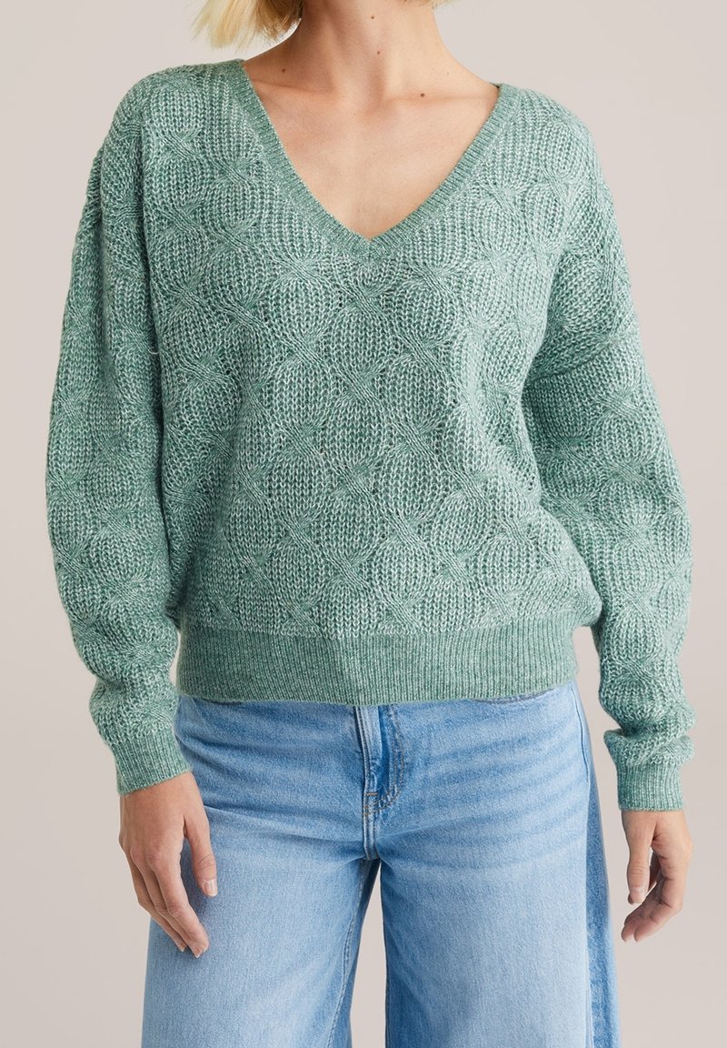 Femme portant un pull en maille texturée vert à col en V avec des manches longues et un jean bleu clair sur un fond uni.