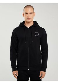 Bărbat purtând un hanorac negru cu fermoar, cu un logo alb circular pe partea stângă a pieptului, pe un fundal simplu de culoare gri deschis.