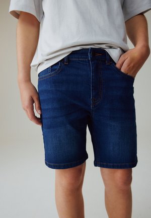 Person trägt dunkelblaue Jeansshorts und ein weißes T-Shirt, eine Hand in der Tasche und die andere seitlich ruhend.