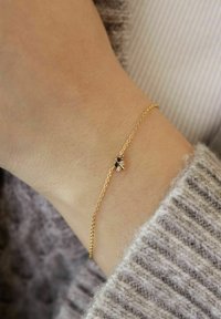 Pulsera de cadena de oro delicada con un pequeño charm de abeja que presenta piedras negras y transparentes, en una muñeca que lleva un suéter de punto gris.
