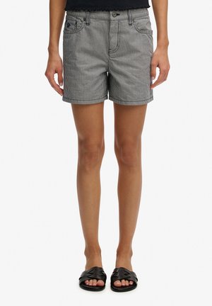 Kvinde iført sort-hvide shorts med lodrette striber og sorte vævede slip-on sandaler, stående mod en hvid baggrund.