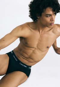 Čierne bavlnené nohavičky s jemným elastickým pásom so značkou "Calvin Klein". Model má svalnatú postavu a kučeravé vlasy.