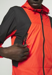 Červená a černá bunda na zip s kapucí, vybavená kapsou na zip na boku, vyrobená z lehké tkaniny s hladkou texturou.