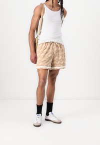 Béžové plavkové šortky s geometrickým vzorem, stahovací šňůrkou v pase a bílým kontrastním lemováním. Nošeny s bílým tílkem a teniskami.