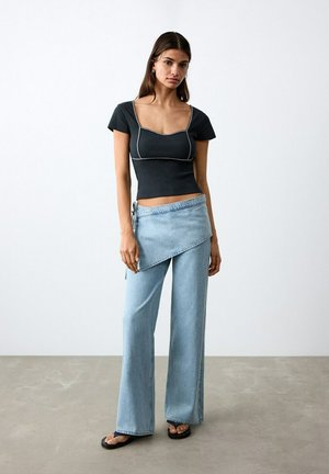 Mlada ženska, ki nosi črn oprijet kratki zgornji del z belim šivanjem, svetlomodre široke kavbojke in črne sandale, stoji ob navadni steni.