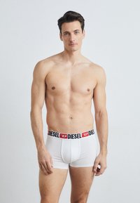 Boxers en coton blancs avec une ceinture logo "DIESEL" noire et rouge. La coupe est ajustée, avec des coutures flatlock. Le modèle pose de manière décontractée.