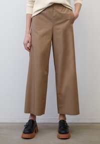 Pantalon marron à jambes larges avec une ceinture élastique et des poches latérales, fabriqué en tissu lisse. Associé à des chaussures noires à plateforme.