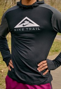Chemise de sport noire à manches longues avec capuche, présentant des accents gris et un logo Nike Trail blanc en forme triangulaire sur la poitrine.