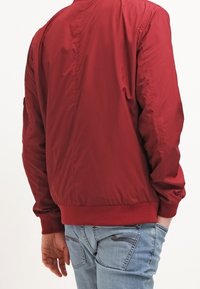 Rote Bomberjacke mit glatter, glänzender Oberfläche, gerippten Bündchen und Saum. Getragen mit hellblauen Jeans aus Denim, die über eine Stichdetails verfügen.