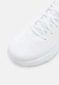 Hvite strikkede løpesko med mesh-overdel, hvite lisser og subtil Under Armour-logo på tungen og nær tåområdet.