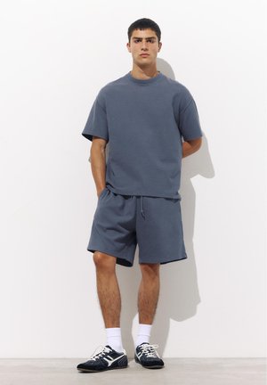 Giovane uomo in piedi con le mani dietro la schiena, indossa una camicia a maniche corte blu abbinata e pantaloncini, calzini bianchi e scarpe da ginnastica nere su sfondo uniforme.