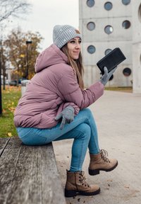 Woman in pink jacket, gray beanie, and gloves sitting on a wooden bench outdoors, holding a black wallet, smiling, autumn setting.