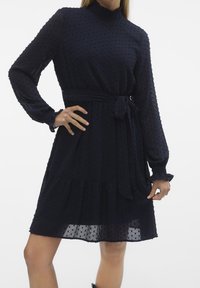 Mörkblå, långärmad klänning med rynkad halsringning, texturerat tyg, midjeband och lager på lager nederkant, som har ett prickigt mönster genomgående.