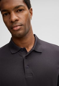 Ciemnoszara koszulka polo wykonana z teksturowanej tkaniny, z plisą na trzy guziki i tradycyjnym kołnierzykiem. Zbliżenie ukazujące dekolt i szczegóły tkaniny.