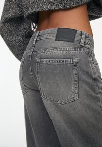 Wytarte szare dżinsy z wysokim stanem, widocznymi kieszeniami i czarną skórzaną łatą na tylnej talii. Teksturowany materiał.