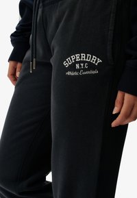 Mørkegrå træningsbukser lavet af blødt stof med et broderet "SUPERDAY N.Y.C Athletic Essentials" logo på venstre lår.