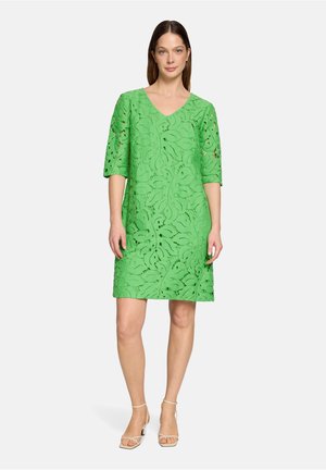 Vrouw draagt een groene kanten jurk tot op de knie met ellebooglange mouwen en beige sandalen met bandjes, staand tegen een effen lichte achtergrond.