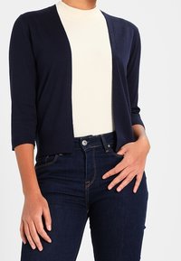 Marinblå kort cardigan med 3/4-långa ärmar, öppen framsida, bärs över en beige ribbad topp, tillsammans med mörka jeans.