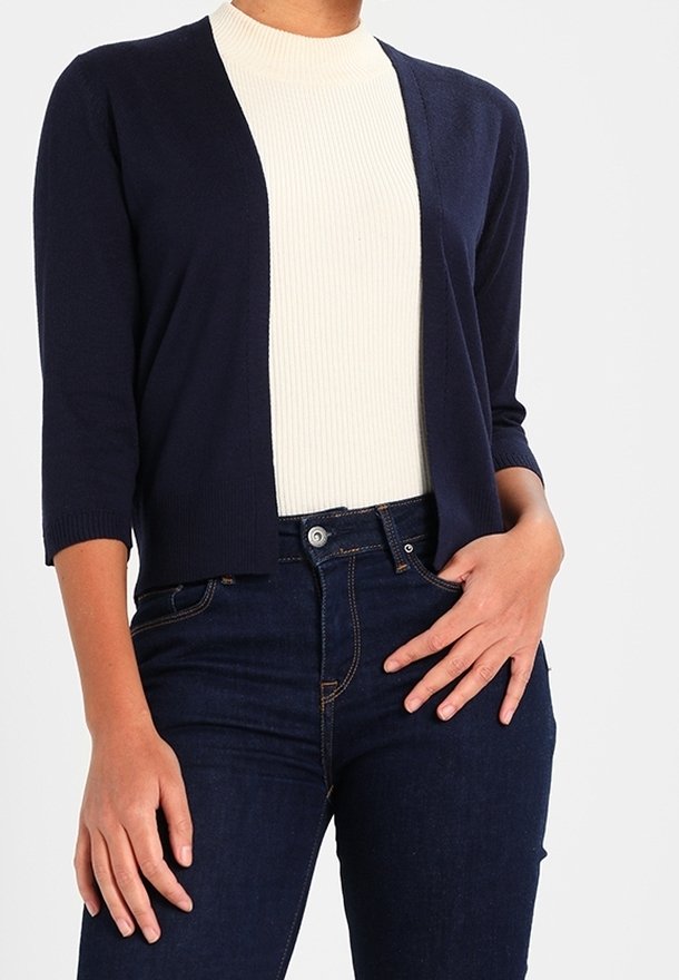 Marinblå kort cardigan med 3/4-långa ärmar, öppen framsida, bärs över en beige ribbad topp, tillsammans med mörka jeans.