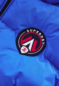 Sinine puffer-jakk, millel on ümmargune tikitud plaastriga "SUPERDRY" tekstiga ning musta, punase ja valge silmapaistva logoga disain.