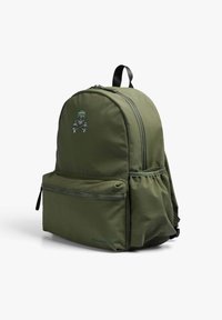 La mochila verde oliva presenta una forma redondeada, compartimentos con cremallera y un bolsillo frontal con un detalle de calavera y tibias bordado.