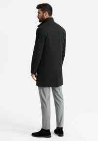 Cappotto in misto lana nero con colletto alto e vestibilità su misura, abbinato a pantaloni grigi e stivali neri alla caviglia. Texture liscia, senza motivi visibili.
