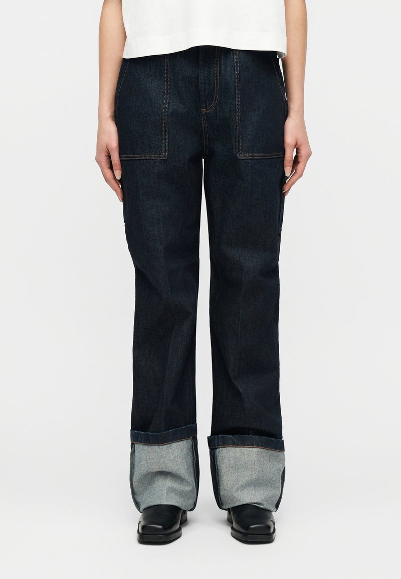 Άτομο που φοράει σκούρο μπλε τζιν με φαρδιά μπατ ζάκια και μεγάλες μανσέτες που δείχνουν το πιο ανοιχτό εσωτερικό ύφασμα, συνδυασμένο με μαύρα παπούτσια και λευκό κοντό τοπ.