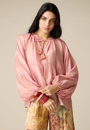 Donna con lunghi capelli neri che indossa una blusa rosa larga con nappine e pantaloni palazzo a fantasia floreale, che tiene al polso braccialetti impilati.