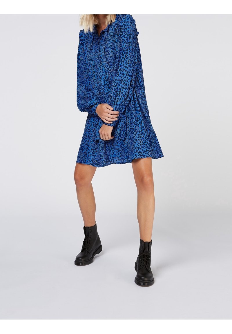 Sininen leopardtäplikkäinen mekko pitkillä hihoilla ja röyhelökauluksella, yhdistettynä mustiin nauhakorkokenkiin. Rakennekangas ja levenevä siluetti.