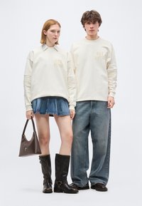 Δύο μοντέλα φορούν ταιριαστά κρεμ φούτερ με το κείμενο «EST 1978», συνδυασμένα με τζιν φούστα και μπότες, καθώς και φαρδιά τζιν με σκούρα παπούτσια.