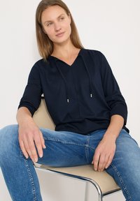 Pull-over bleu marine avec col en V, capuche à cordon et manches trois quarts. Associé à un jean en denim bleu clair. Texture lisse, coupe décontractée.