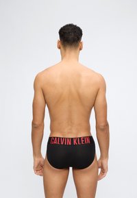 Άνδρας που στέκεται με την πλάτη στην κάμερα φορώντας μαύρα εσώρουχα Calvin Klein με κόκκινο λογότυπο στη ζώνη μέσης, σε απλό λευκό φόντο.