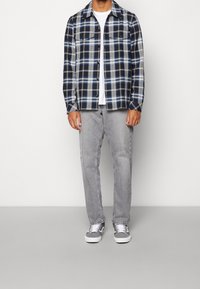 Niebieska i czarna koszula flanelowa w kratę na białym t-shircie, skomponowana z szarych dżinsów i szarych sneakersów. Posiada dwie kieszenie na piersi.