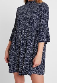 Granatowa sukienka w białe plamki, z wysokim kołnierzem i rozszerzanymi rękawami. Charakteryzuje się warstwowym krojem oraz miękką, lekką tkaniną.