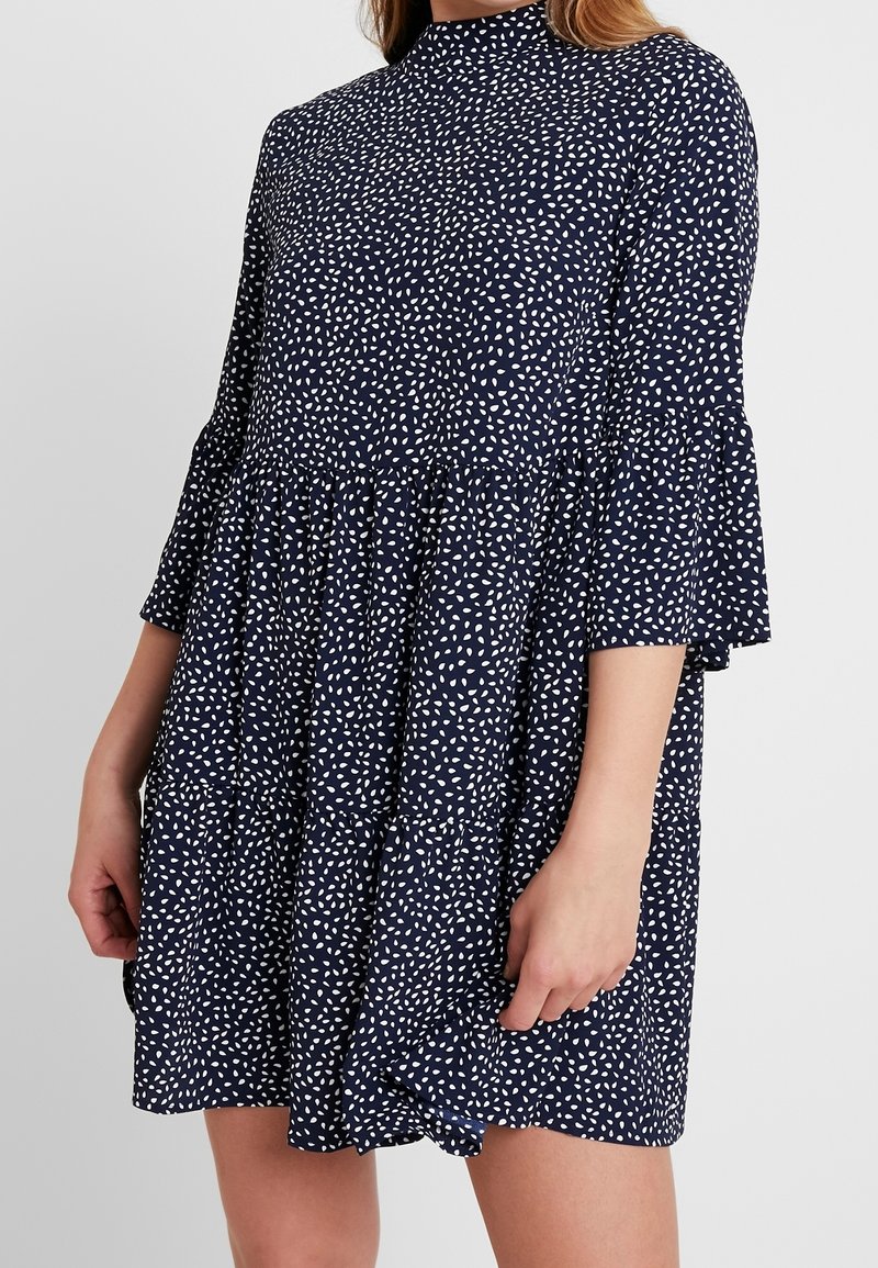 Granatowa sukienka w białe plamki, z wysokim kołnierzem i rozszerzanymi rękawami. Charakteryzuje się warstwowym krojem oraz miękką, lekką tkaniną.