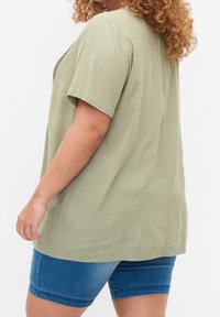 Olive green short-sleeved blouse with a loose fit, textured fabric, and a rounded neckline, paired with blue denim shorts.