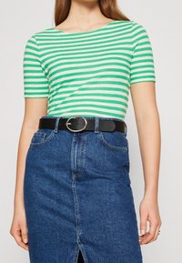 Nainen, jolla on päällään vihreä-valkoisia raitoja sisältävä lyhythihainen paita, joka on täytetty sinisiin denimitaskuhousuihin, joissa on musta vyö, jossa on hopeinen pyöreä solki.