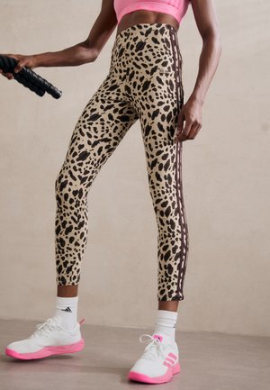Legginsy w wzór lamparta w beżowym kolorze z czarnymi plamami, z brązowymi paskami po bokach i elastycznym pasem. Połączone z białymi sneakersami z różowymi akcentami.