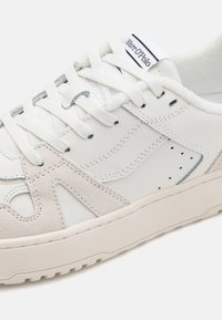 Weiße LederSneaker mit beigen Wildleder-Akzenten, strukturierten Überzügen und durchbrochenen Details. Verfügbar mit weißen Schnürsenkeln und einer schlichten Gummisohle.