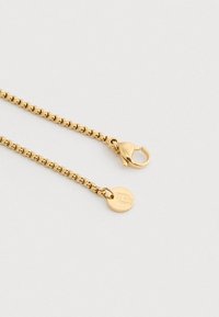 Χρυσή αλυσίδα κολιέ με κούμπωμα αχίλλειου και μία μικρή στρογγυλή ετικέτα με χαραγμένο ένα στυλιζαρισμένο λογότυπο, τοποθετημένη σε ουδέτερο φόντο.