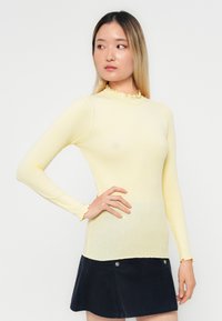 Langarm geribbeld top in lichtgeel met een rimpelmocknek en manchetten, gecombineerd met een marineblauwe corduroy rok.