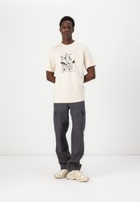 Beige bomulds t-shirt med grafisk print i sort, parret med mørkegrå cargo-bukser med flere lommer og elastiske manchetter.