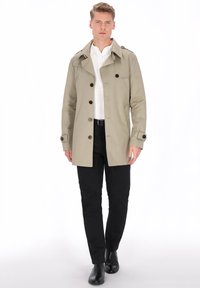 Gabardina beige con botones decorativos, cuello estructurado y bolsillos laterales, llevada sobre una camisa blanca y pantalones negros, combinada con botas negras.