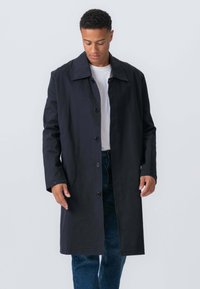 Mand iført en lang sort knappet frakke over en hvid skjorte og blå jeans, som ser ned mod en ensfarvet grå baggrund.