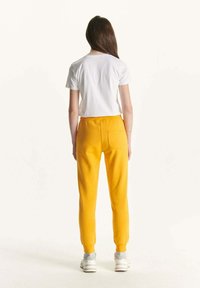 Geel trainingsbroek met een gladde textuur en elastische boorden, gecombineerd met een eenvoudige witte T-shirt. Bevat achterzakken en heeft een relaxte pasvorm.