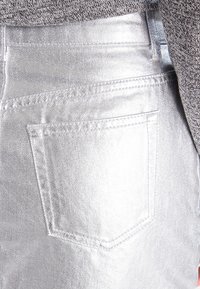 Metalické stříbrné džíny s kapsou na zadní straně, hladkou texturou a viditelnými švy. Pas je částečně viditelný.