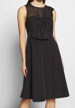 Sort ærmeløs kjole med gennemsigtigt øverste brystpanel og stort stofsløjfedetalje foran, båret af en stående person.