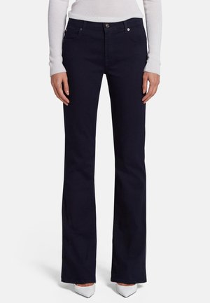 Kobieta ubrana w czarne jeansy bootcut, białe szpilki z ostrym noskiem i biały prążkowany top z długim rękawem, stojąca na jednolitym tle.