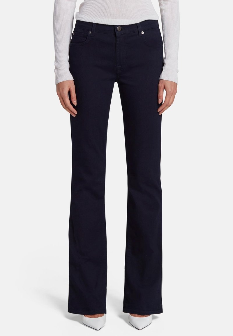 Femme portant un jean bootcut noir, des talons blancs à bout pointu et un haut blanc à manches longues côtelé, debout devant un fond uni.