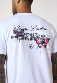 Weißes Baumwoll-T-Shirt mit einem grafischen Design von lila und rosa tropfenden Schmetterlingen sowie dem Text: "Closure London" und "Creators of Modern Essentials."
